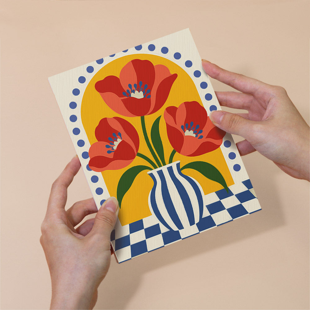 A pair of hands holding a card with a vibrant illustration of red flowers in a vase against a checkered background.