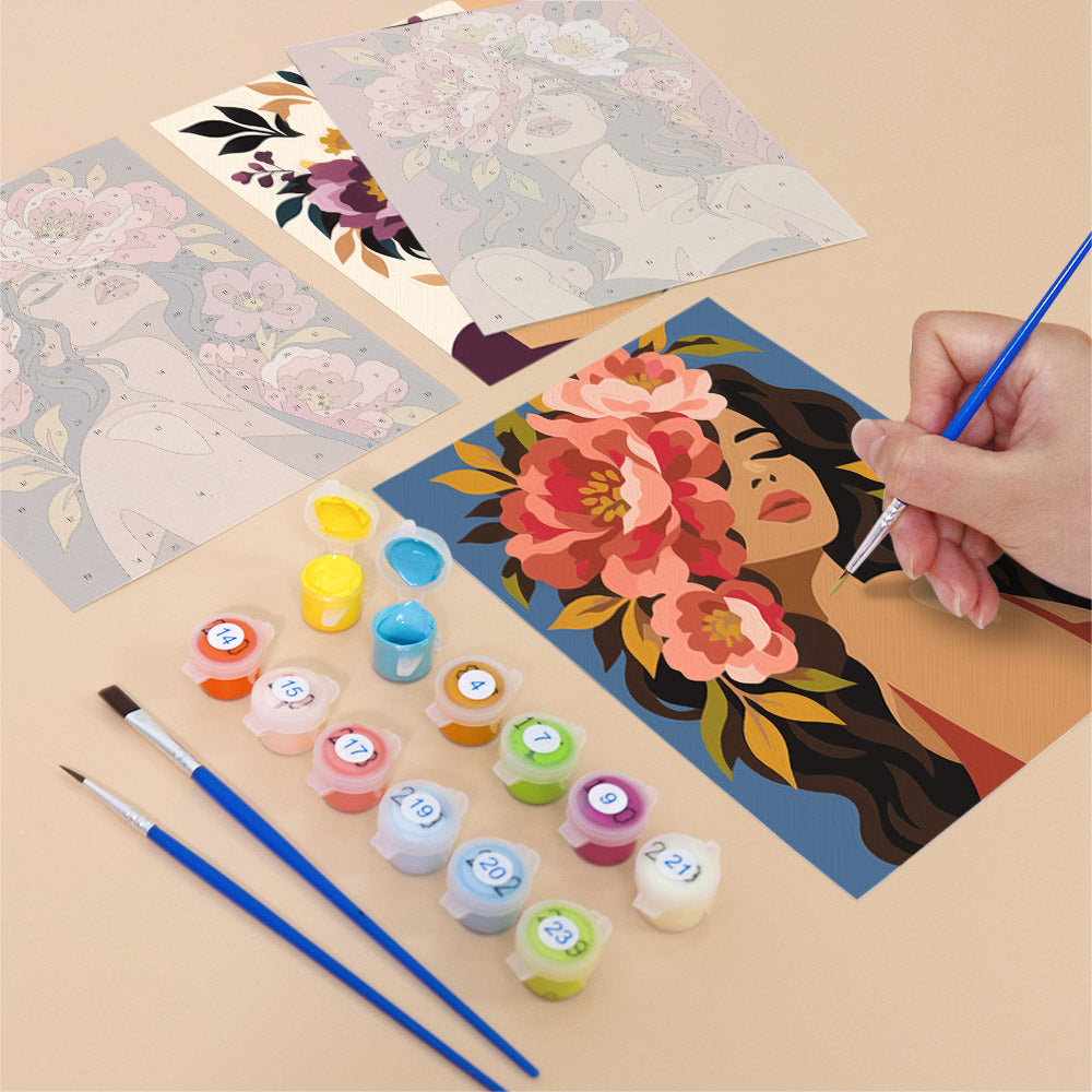 a person's hand holding a paintbrush, surrounded by art supplies such as paint tubes and brushes, as well as a partially completed painting featuring a woman's face with flowers.