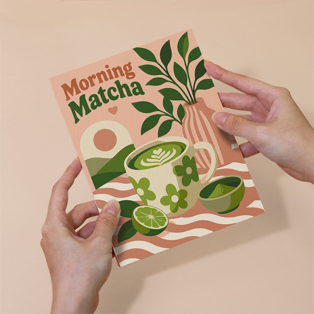 A person's hands holding a book or magazine with a colorful illustration of a cup of matcha tea, a plant, and other green elements on a light peach background.