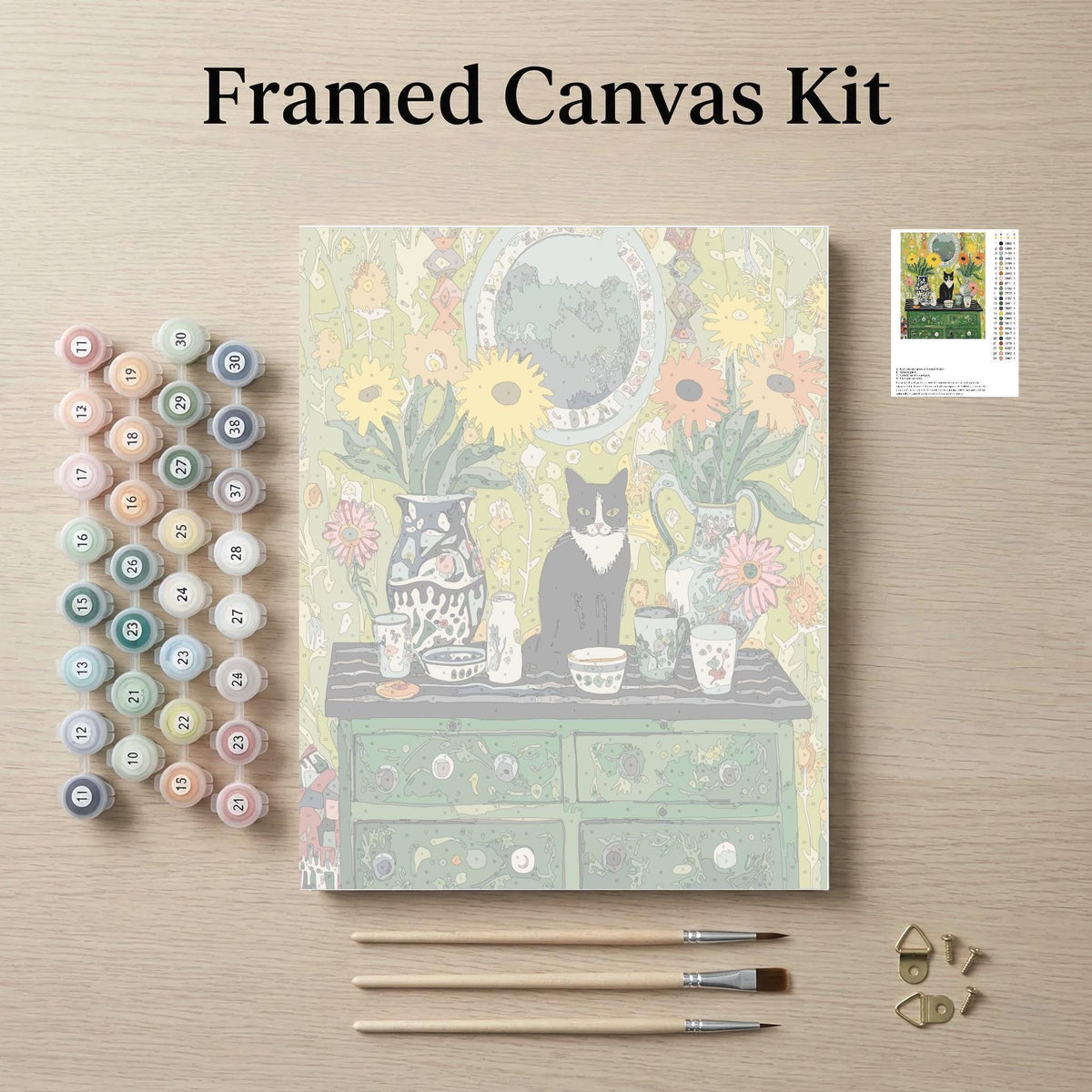 The image is a framed canvas kit featuring a painting of a cat sitting on a table surrounded by various objects, such as vases, cups, and a clock. The kit also includes paintbrushes, paint tubes, and a small picture frame.
