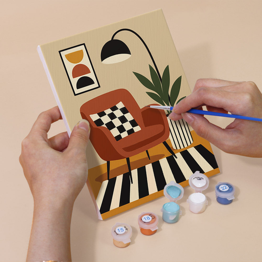 A person's hands are holding a painting kit with a checkered chair and a potted plant, and there are paint tubes and a paintbrush nearby, suggesting that the person is about to start painting a picture of the chair and plant.