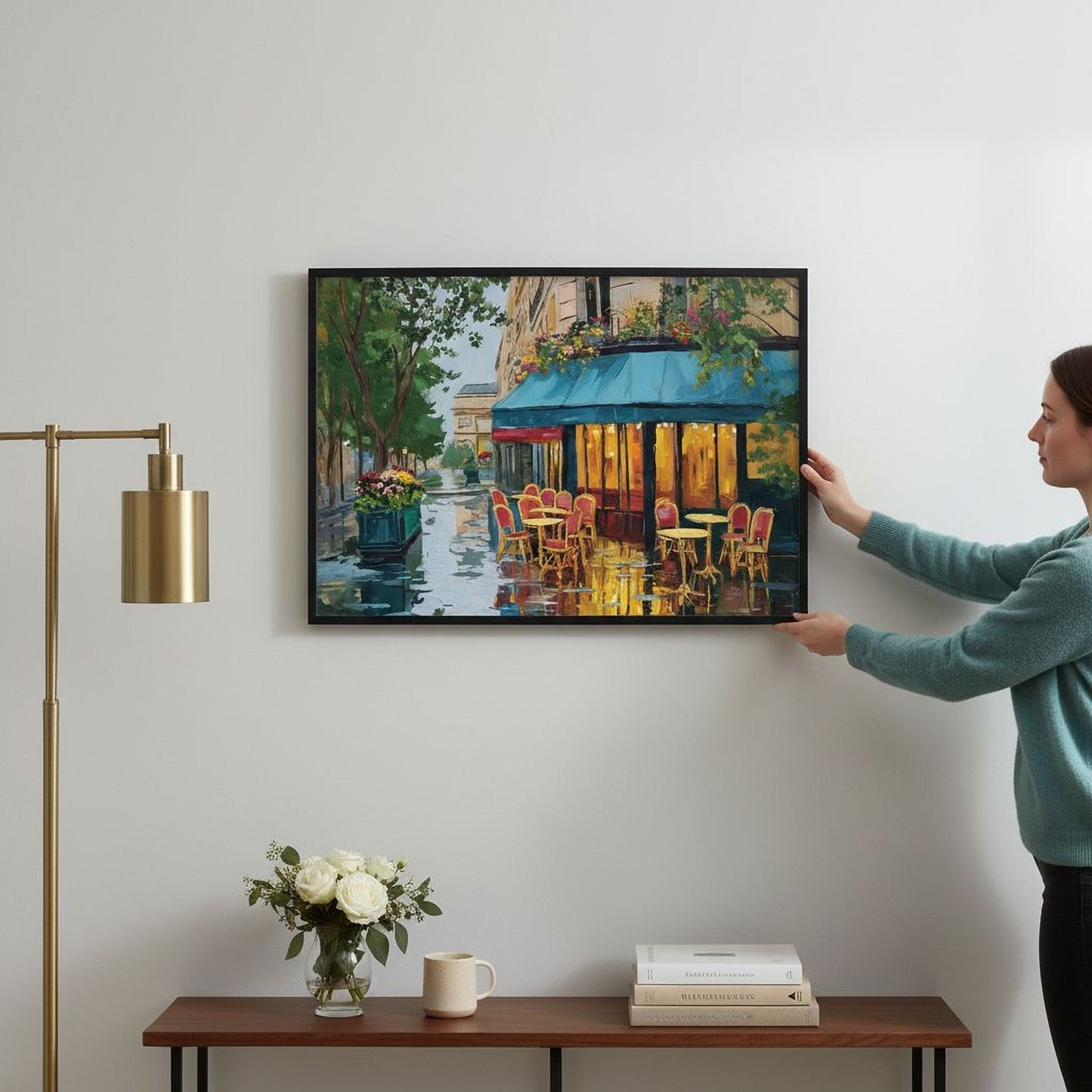 A woman is hanging a framed painting of a colorful street scene on a wall, which features a cafe with outdoor seating and a fountain.