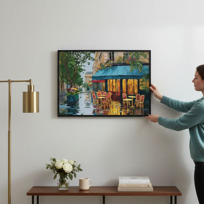 A woman is hanging a framed painting of a colorful street scene on a wall, which features a cafe with outdoor seating and a fountain.