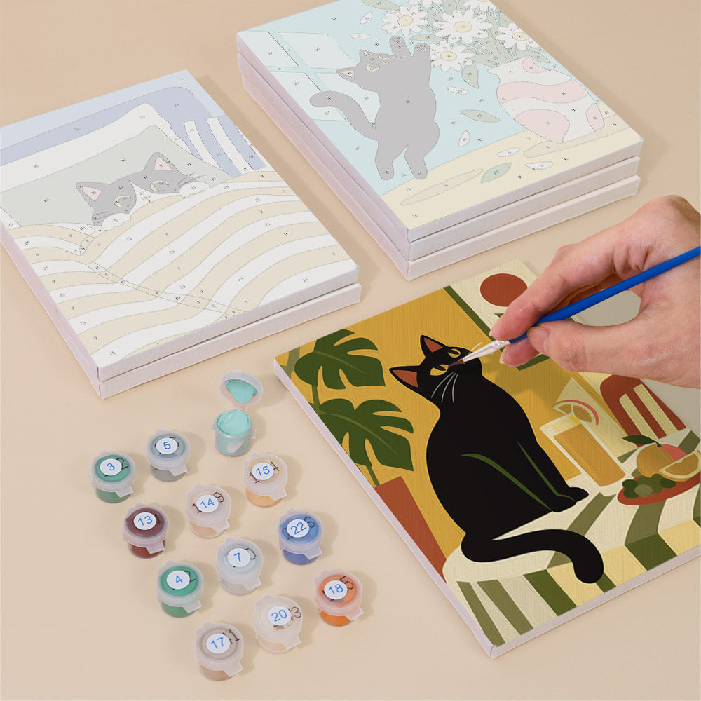a person's hand painting a picture of a black cat on a canvas, surrounded by various art supplies such as paint bottles and a sketchbook.