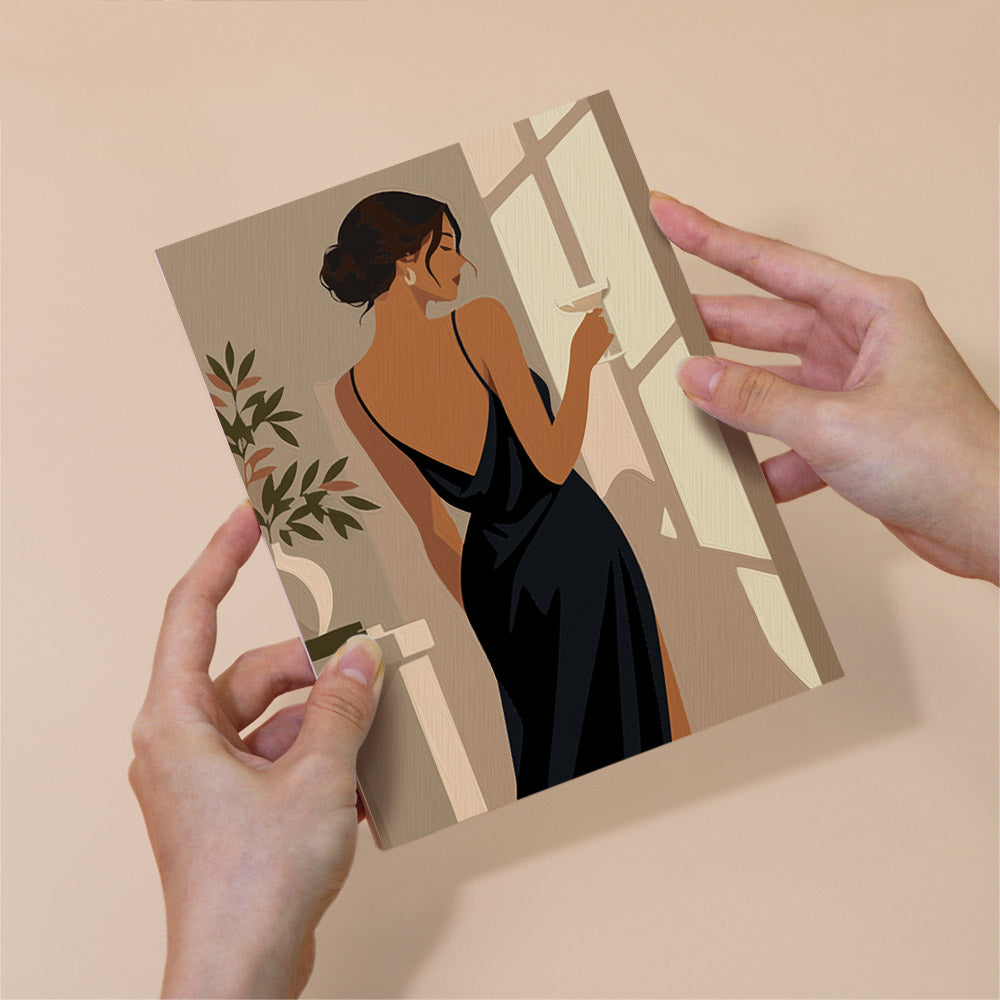 A person's hands holding a framed illustration of a woman in a black dress, with a potted plant and a window in the background.
