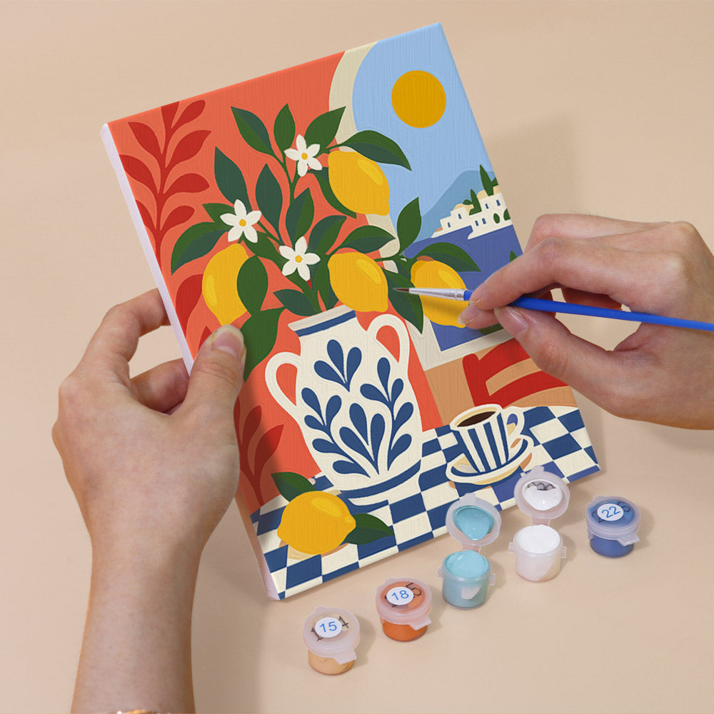 A person's hands are holding a painting kit with a vibrant, colorful painting depicting a vase with flowers and a lemon tree. The kit includes paint colors and brushes, and there are additional paint colors and brushes on the table.