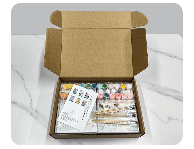 An open cardboard box containing various art supplies, including paint tubes, brushes, and a guidebook, placed on a marble countertop.