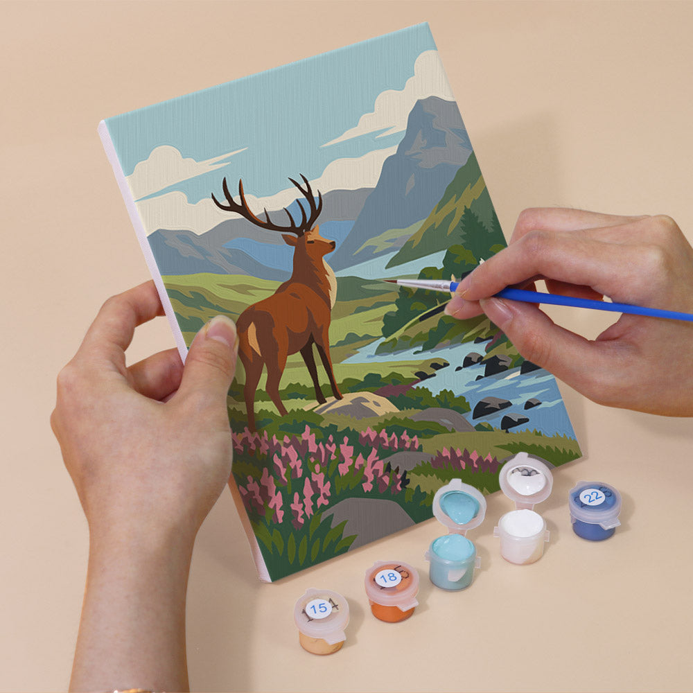 A person's hands are holding a painting kit with a landscape scene featuring a deer, mountains, and a river. The kit includes paint colors and brushes, and there are paint containers with numbers on them, likely indicating the colors used in the painting.