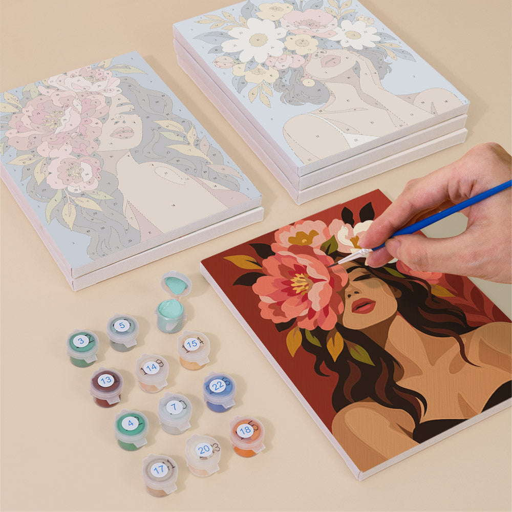 a person's hand painting a floral portrait on a canvas, surrounded by art supplies such as paint bottles and a sketchbook.