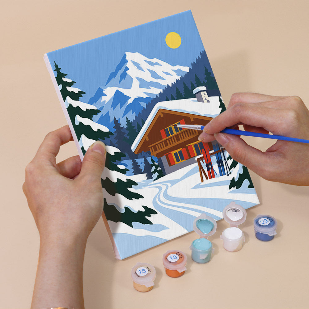 A person's hands are holding a painting kit with a winter landscape depicting a cabin on a snowy mountain, surrounded by pine trees.