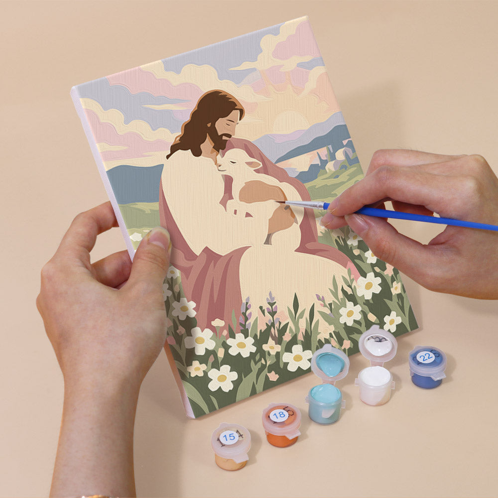 A person's hands are holding a painting kit with a canvas depicting a religious scene, and there are paint bottles and brushes nearby, suggesting that the person is in the process of painting the scene.