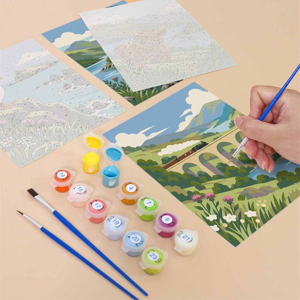 a person's hand holding a paintbrush, surrounded by art supplies and a painting depicting a scenic landscape with a bridge and flowers.