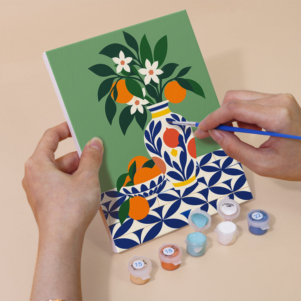 A person's hands are holding a painting kit with a green canvas featuring a vase with flowers and oranges, and a blue and white patterned background.