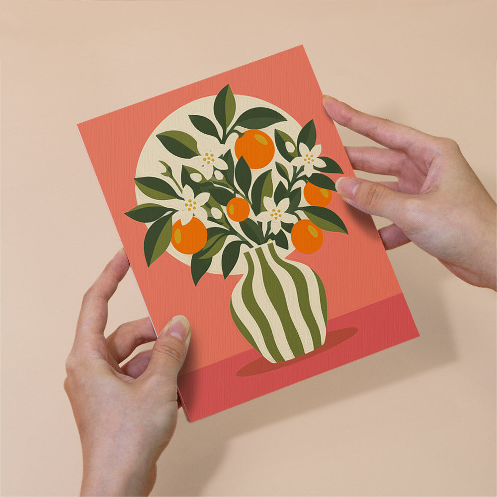 A pair of hands holding a card with a vibrant illustration of a vase filled with orange flowers and green leaves against a pink background.