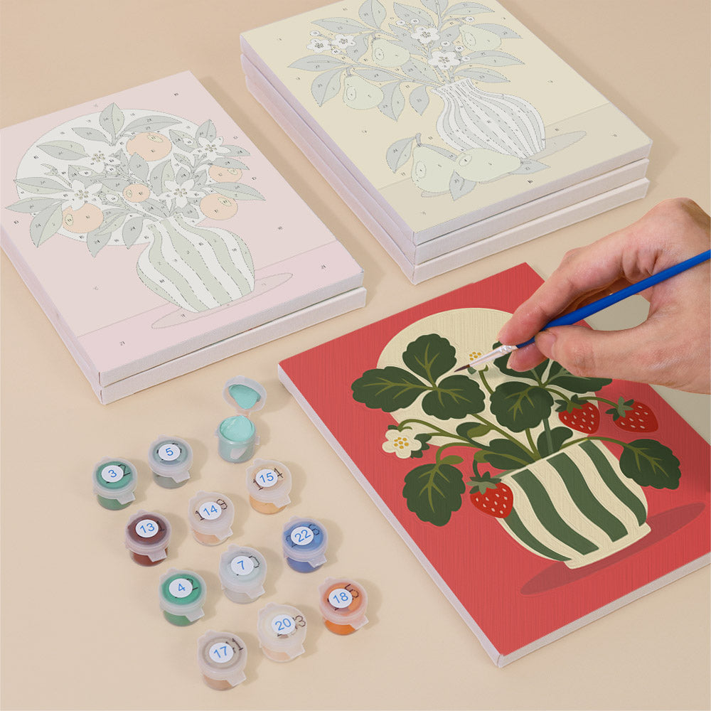a person's hand painting a strawberry plant on a red canvas, surrounded by various art supplies such as paint bottles and a sketchbook.