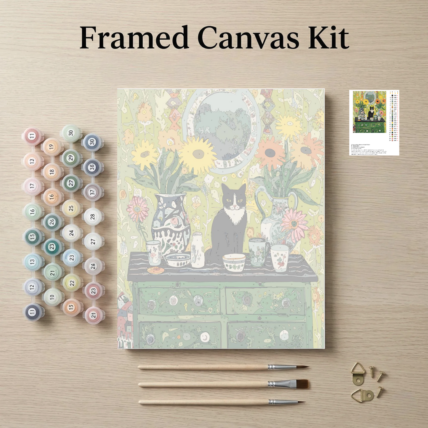 The image is a framed canvas kit featuring a painting of a cat sitting on a table surrounded by various objects, such as vases, cups, and a clock. The kit also includes paintbrushes, paint tubes, and a small picture frame.