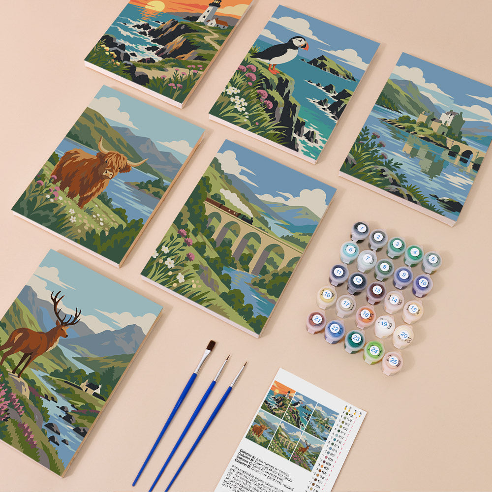 six illustrated paintings depicting various landscapes, including mountains, a lighthouse, a castle, a bridge, and a moose. There are also paintbrushes and paint tubes arranged on a table, suggesting that this is an art kit for painting these landscapes.