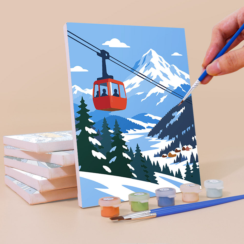 A hand is painting a picture of a ski lift carrying passengers over a snowy mountain landscape, with a set of paintbrushes and paint tubes nearby.