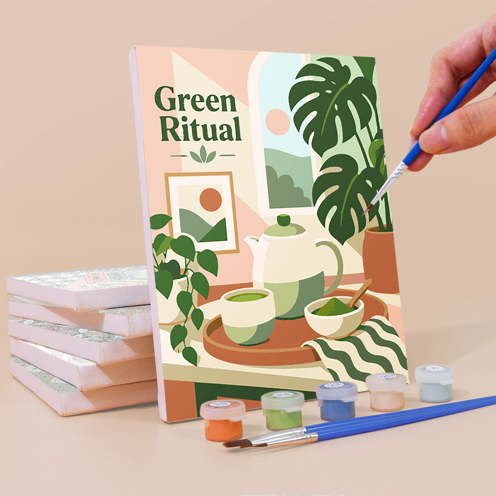 a hand holding a paintbrush, with a stack of books and art supplies in the foreground, and a painting of a kitchen scene with a teapot, bowls, and plants on the cover of a book titled "Green Ritual".