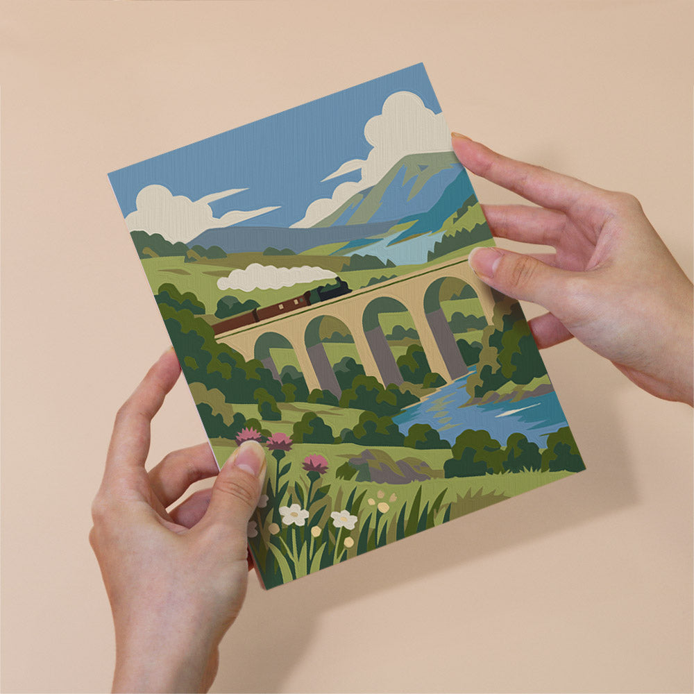 A person's hands holding a card with a colorful illustration of a train passing under a bridge over a river, surrounded by lush greenery and flowers.