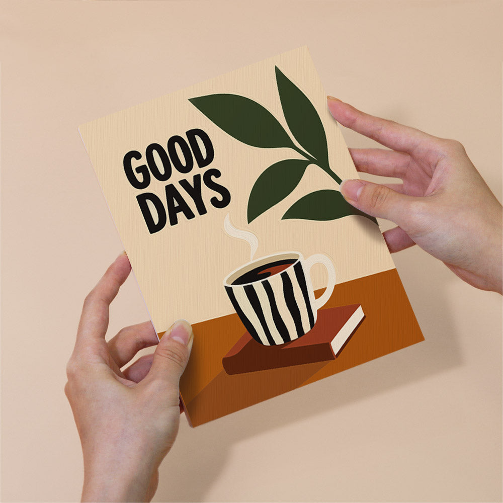 A person's hands holding a card with the text "GOOD DAYS" and an illustration of a striped coffee mug on a book.