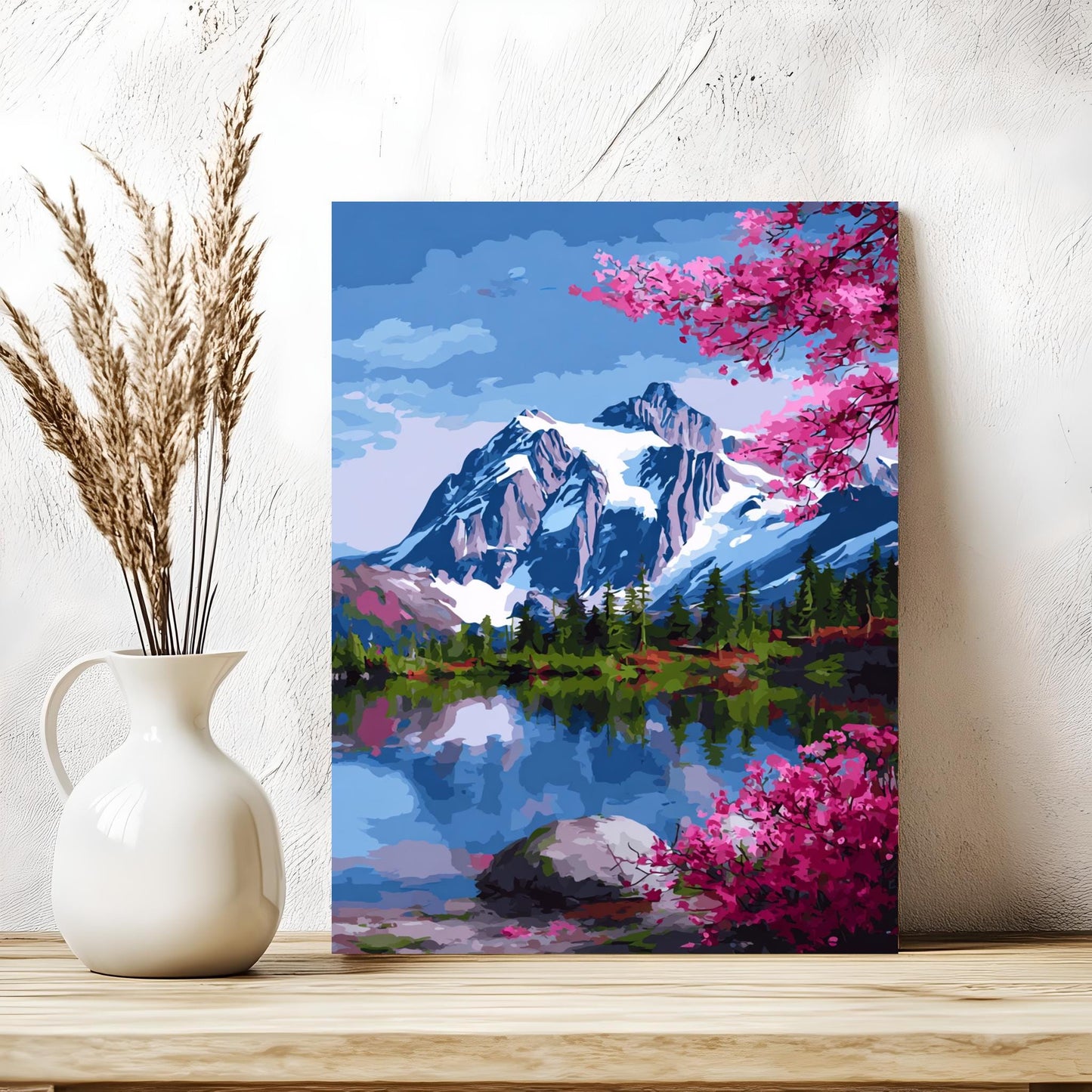 a painting of a mountain landscape with a lake and pink flowers, displayed on a wooden shelf next to a white vase with dried reeds.
