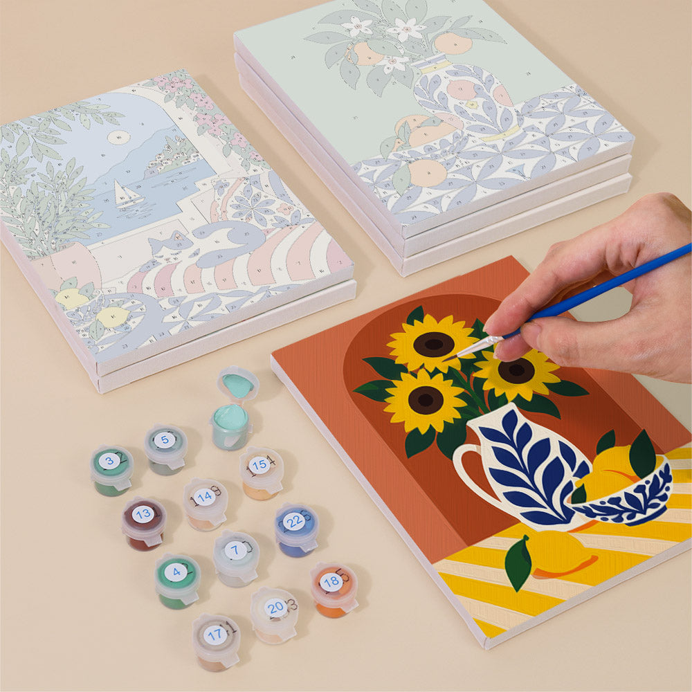 a person's hand painting a picture of a vase with flowers on a piece of paper, surrounded by various art supplies such as paint tubes and a sketchbook.