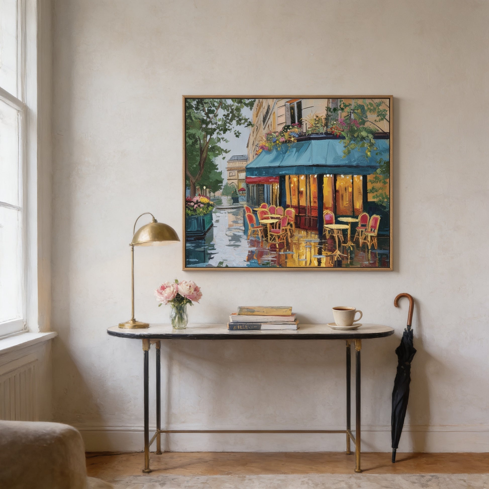 a painting of a Parisian street scene hanging on a wall, with a table and chairs below it. The table has a vase of flowers, a lamp, and a book on it. There is also an umbrella leaning against the wall.