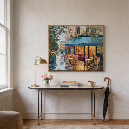 a painting of a Parisian street scene hanging on a wall, with a table and chairs below it. The table has a vase of flowers, a lamp, and a book on it. There is also an umbrella leaning against the wall.