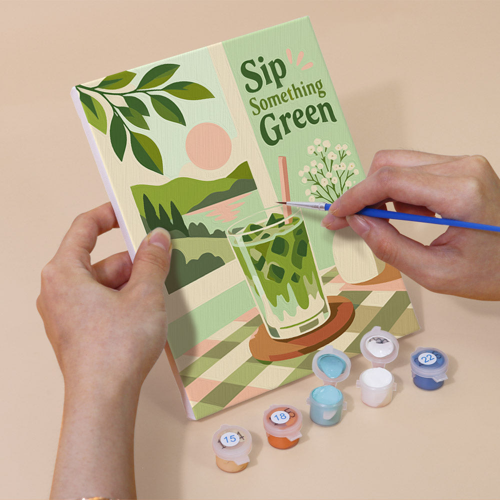 A person's hands holding a book with a painting of a green drink on the cover, along with paint supplies and a brush.