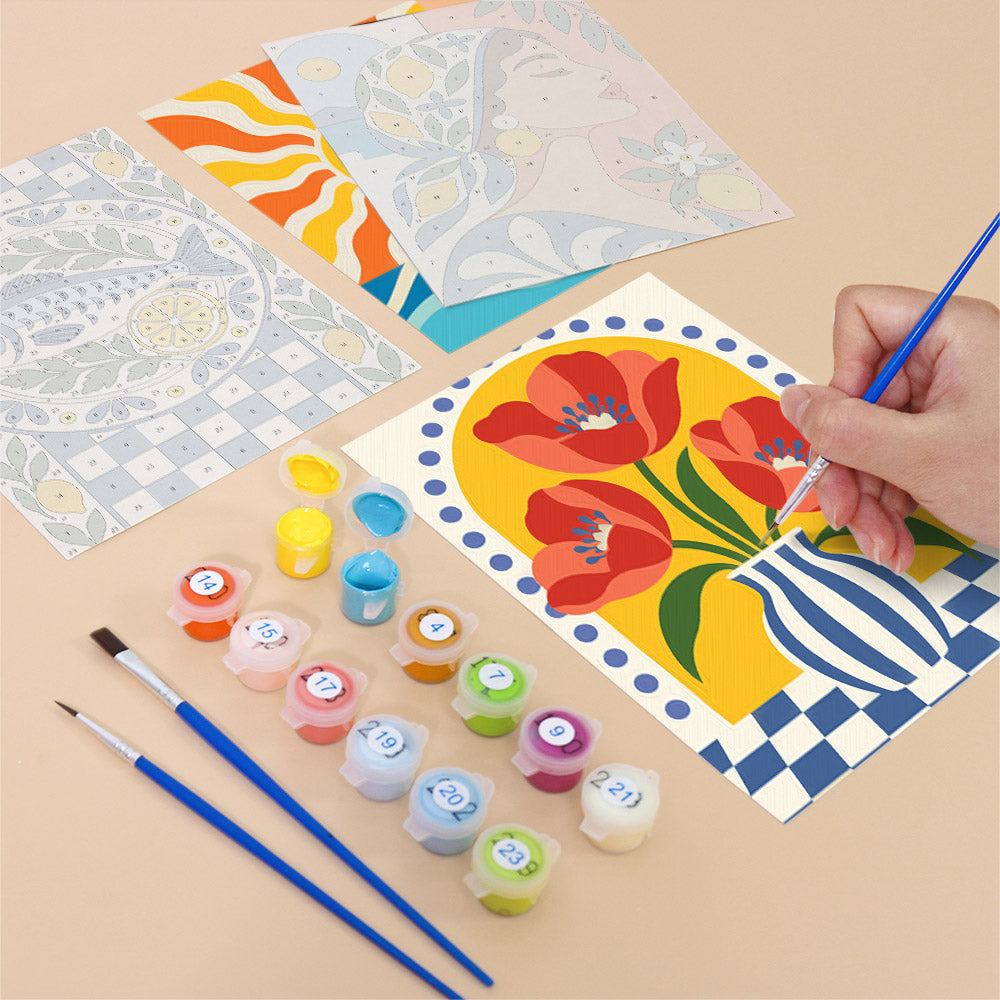 a person's hand holding a paintbrush, painting a picture of a vase with red flowers on a yellow background. Various art supplies, including paint tubes and brushes, are scattered around the workspace.