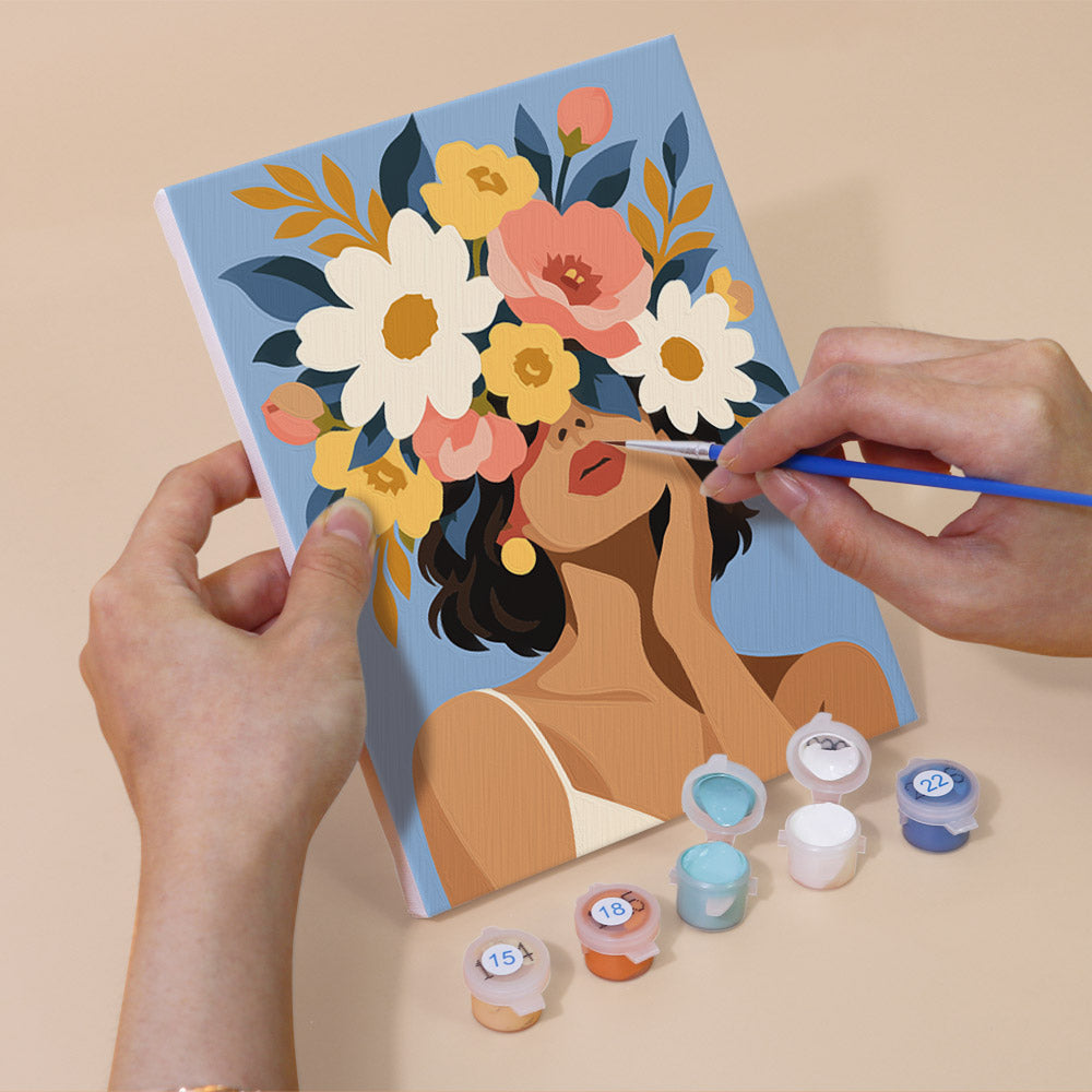 A person's hands are holding a canvas with a floral design, and there are paint bottles and a paintbrush nearby, suggesting that the image is a painting kit.