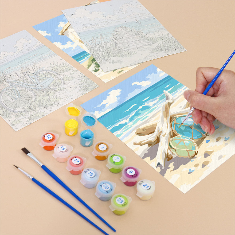 a person's hand painting a picture of a beach scene using watercolor paints and brushes. There are several paint tubes and brushes arranged on a table, along with a completed painting depicting a beach scene.