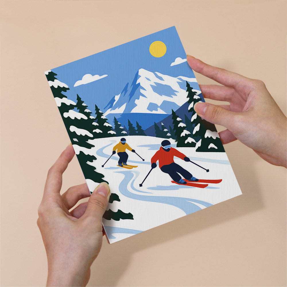 A pair of hands holding a card with a winter scene depicting two skiers on a snowy mountain slope.