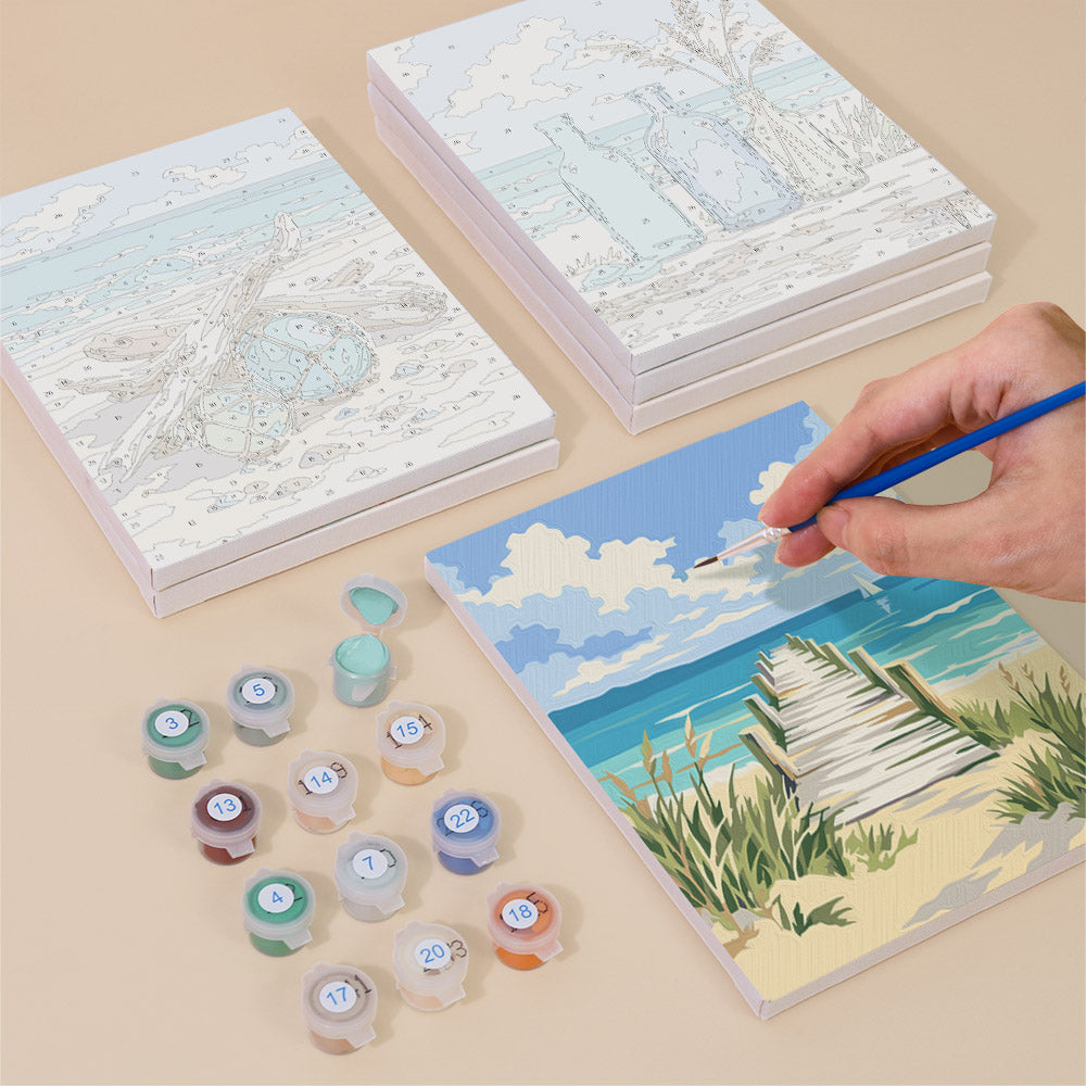 a person's hand painting a picture of a beach scene using a paintbrush and a palette of colors. There are also several paint cans and a sketchbook with a beach scene illustration on it, suggesting that the person is engaged in an artistic activity.