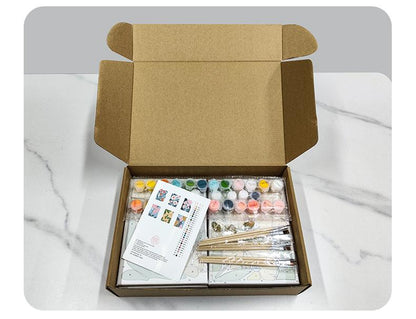 An open cardboard box containing various art supplies, including paint tubes, brushes, and a guidebook, placed on a marble countertop.