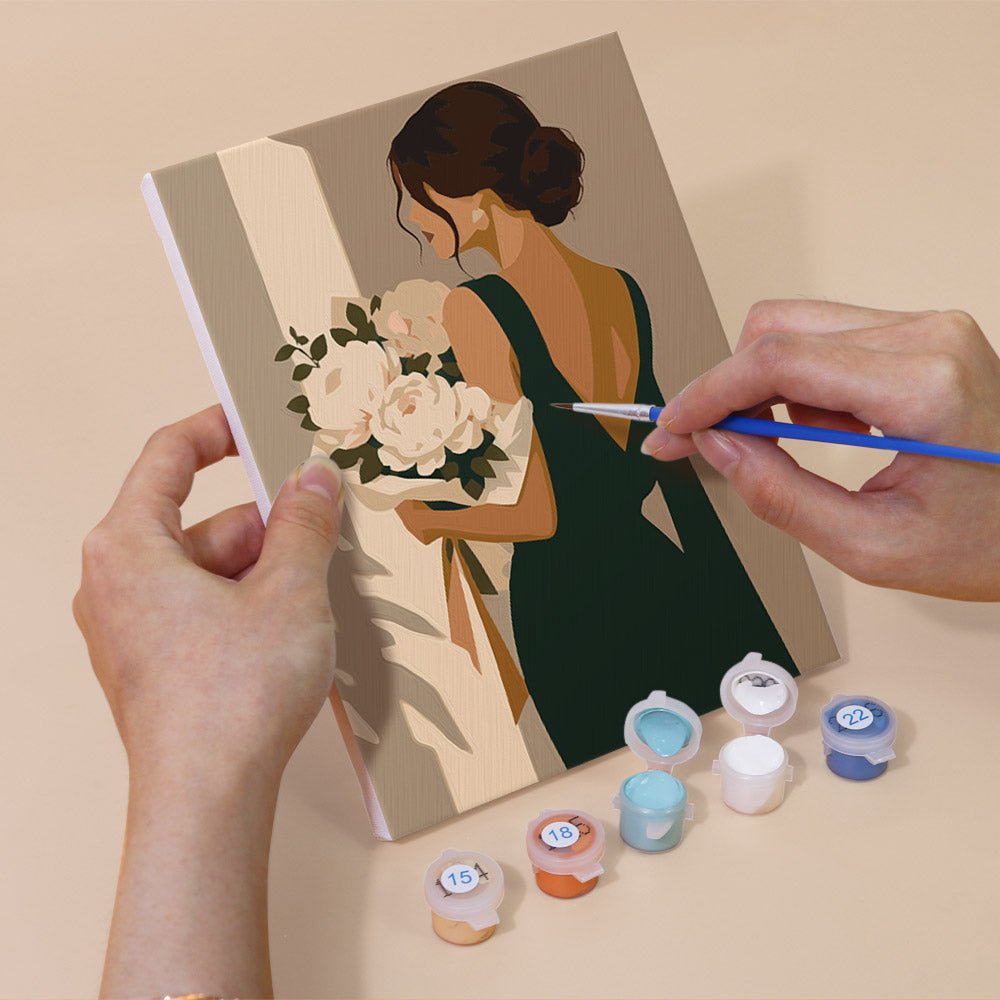 A person's hands are holding a painting of a woman holding a bouquet of flowers, with paintbrushes and paint tubes in front of the artwork.
