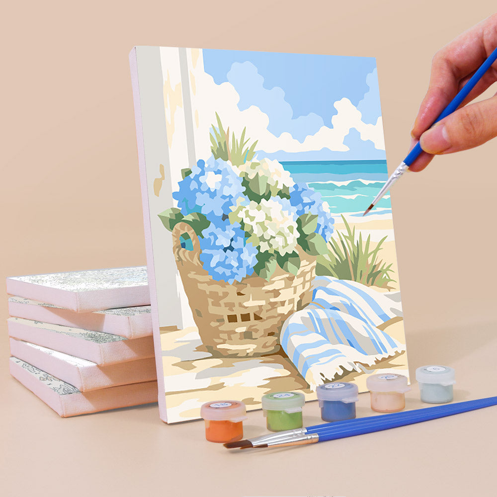 a painting of a basket filled with blue and white flowers, placed on a table with a paintbrush and a set of paint colors.