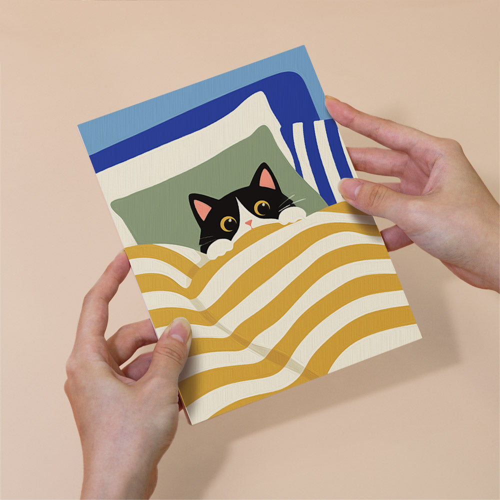 A person's hands are holding a card with a black and white cat peeking out from under a striped blanket.