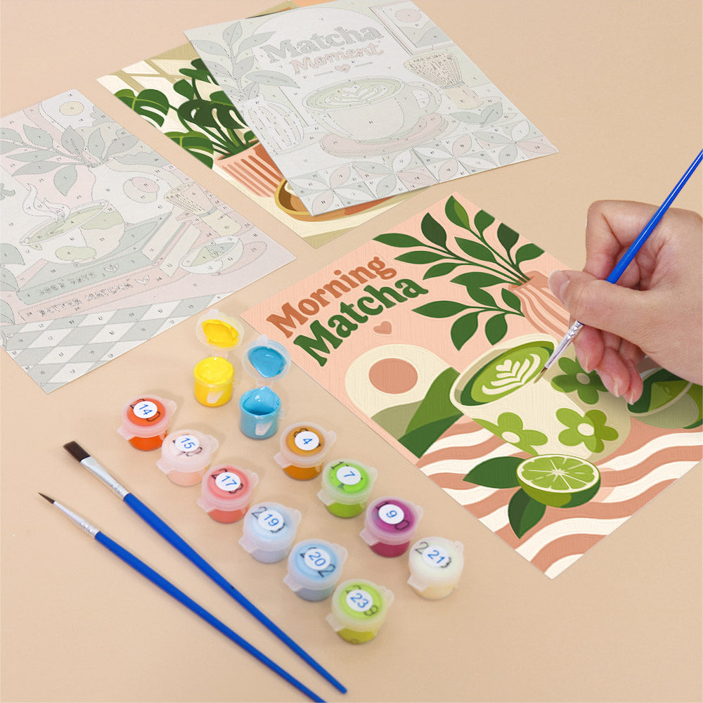 a hand painting a picture of a cup of matcha, surrounded by art supplies such as paintbrushes and paint tubes.