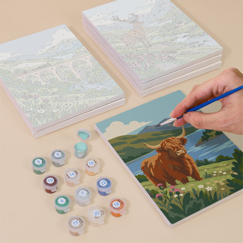 a person's hand holding a paintbrush, painting a picture of a brown cow in a grassy field with a river in the background. There are also several paint color bottles and a set of blank canvas sheets, suggesting that the person is engaged in an art project.