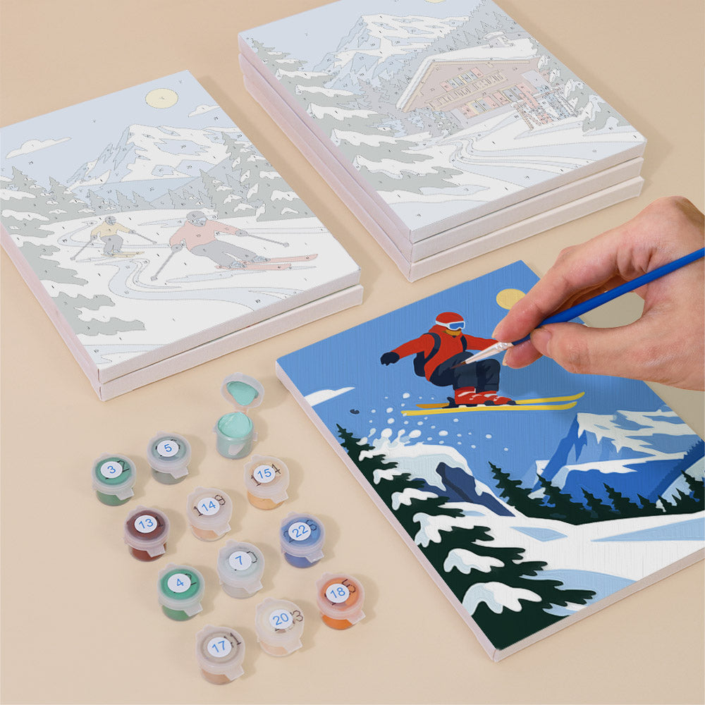 a person's hand painting a picture of a skier on a snowy mountain, surrounded by art supplies and a set of books with illustrations of snowy mountain scenes.
