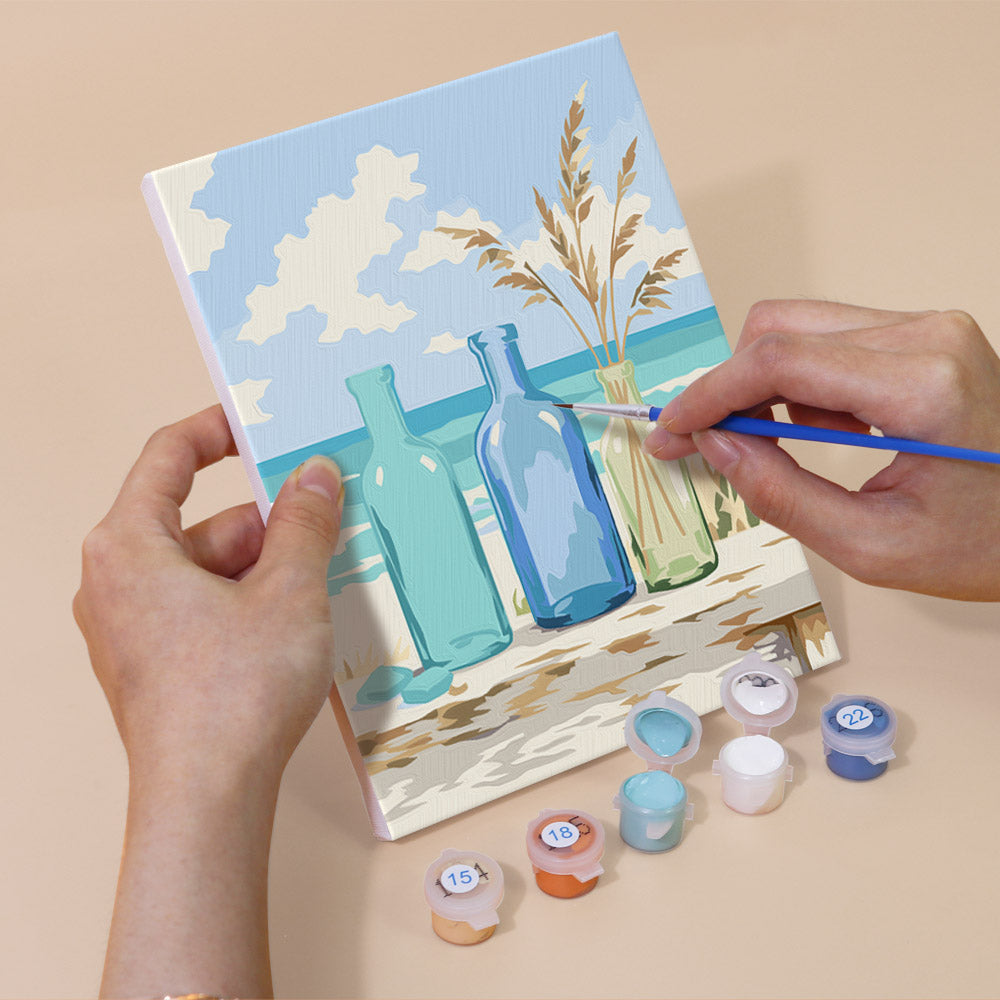 A person's hands are holding a painting kit with a canvas featuring a beach scene with two bottles and a vase, along with various paint colors and brushes.