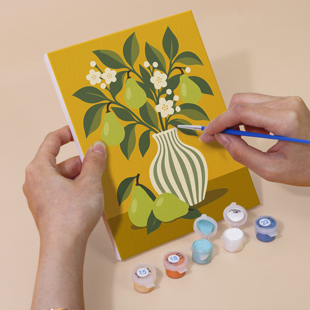 A person's hands are holding a painting kit with a yellow canvas featuring a vase with green pears and white flowers. The kit also includes paint colors and a paintbrush.