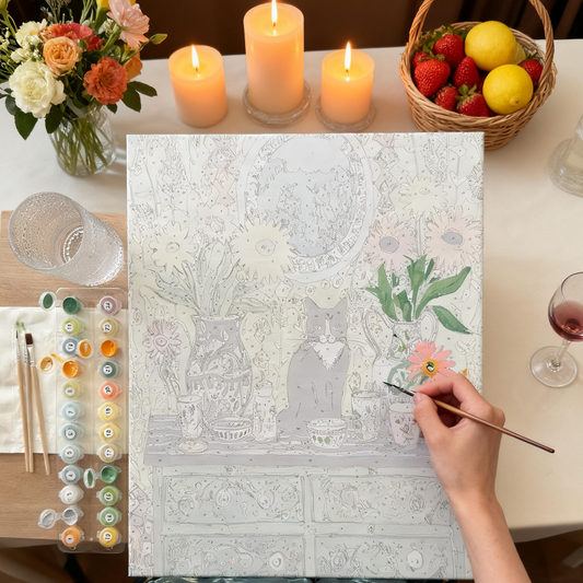 a person's hand painting a picture of a cat sitting on a table with various objects, such as vases, flowers, and a bowl. The painting is surrounded by art supplies, including paintbrushes and a palette of colors.