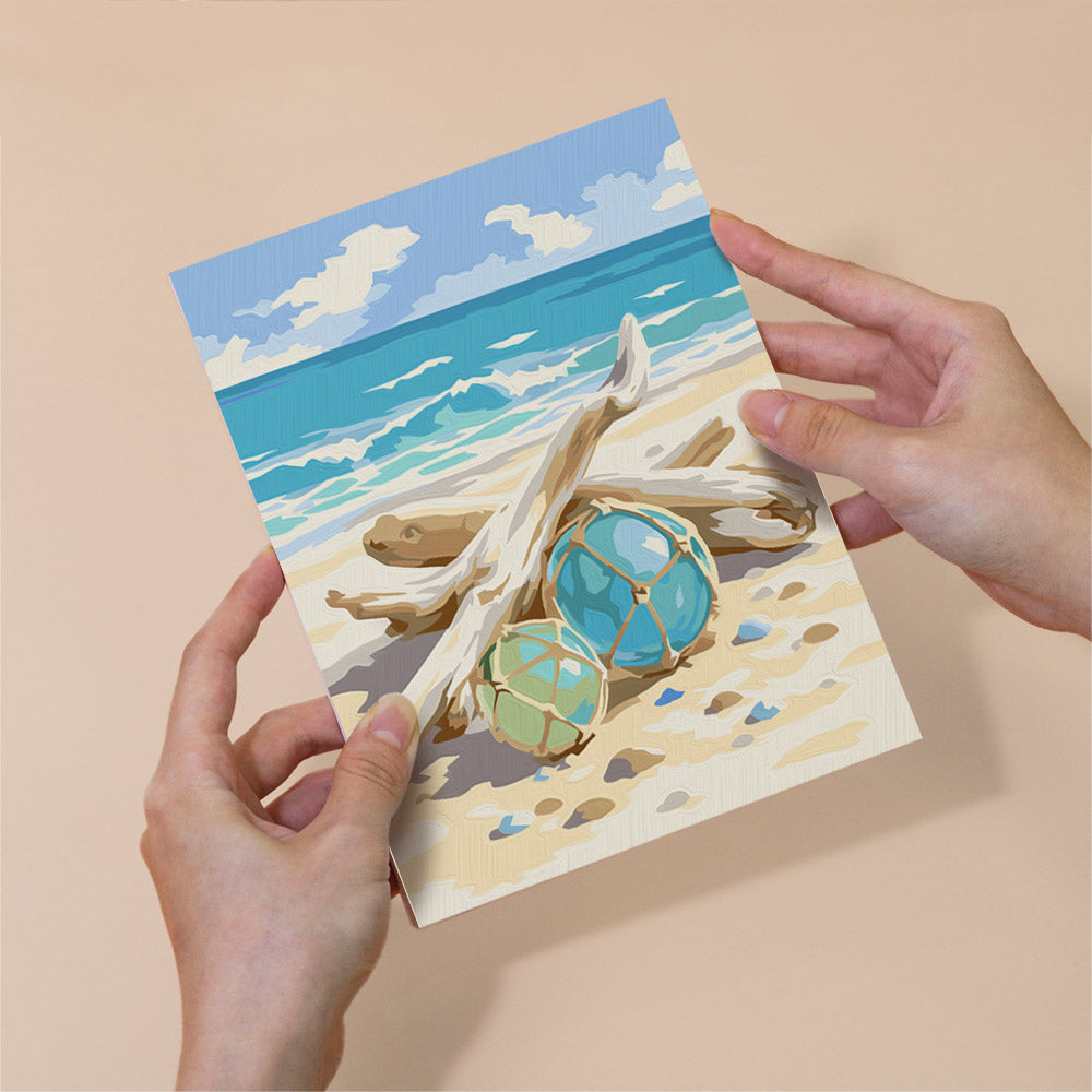 A person's hands holding a card with a beach scene featuring driftwood, blue and green objects, and a cloudy sky.