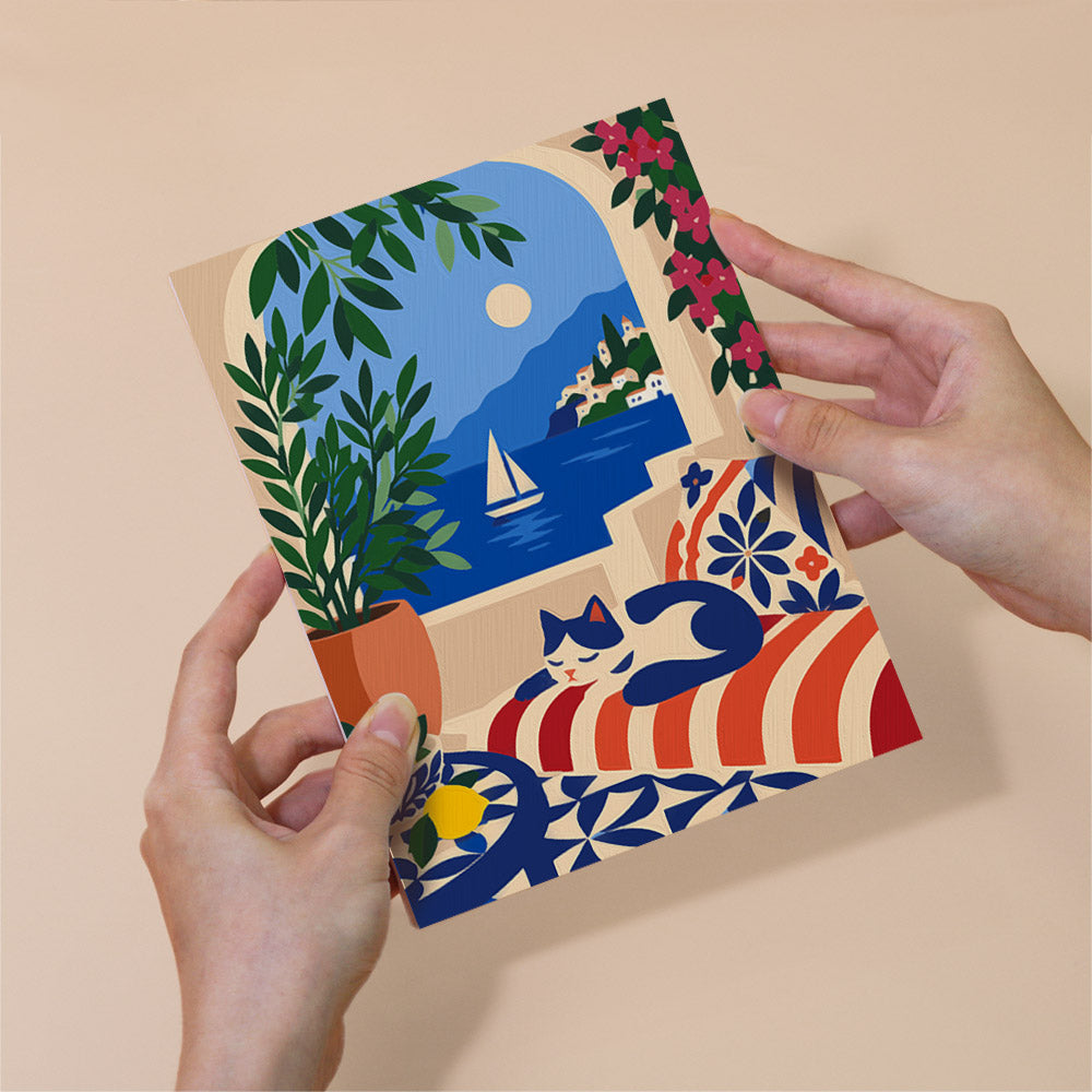 A person's hands holding a colorful illustration depicting a serene coastal scene with a sailboat, a cat, and a potted plant.